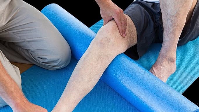 Fysiotherapeut begeleidt een patiënt bij beenrevalidatie met foamroller op de mat